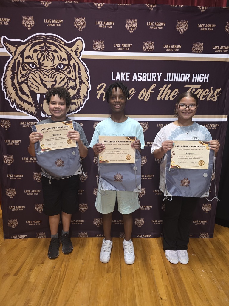 Three children hold certificates in front of a sign with a tiger and school name.