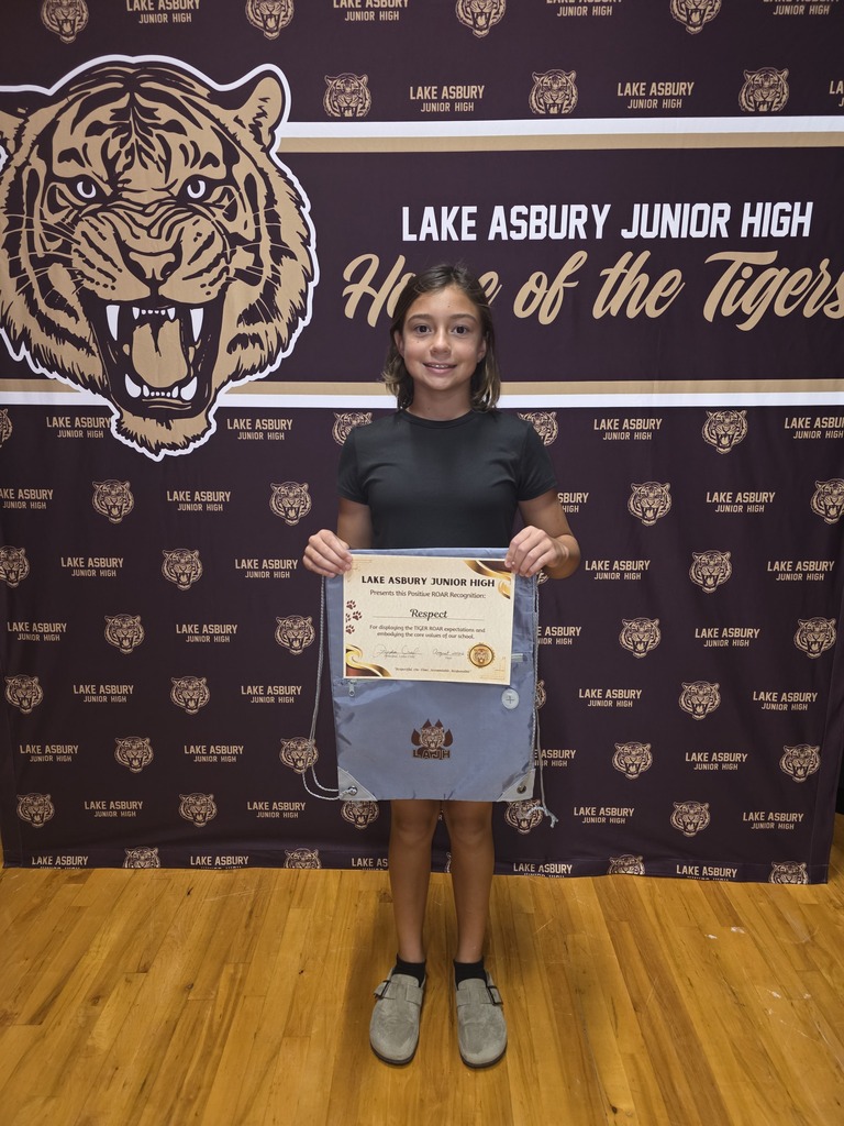 A young girl holds a certificate in a room with a large tiger mascot on the wall.
