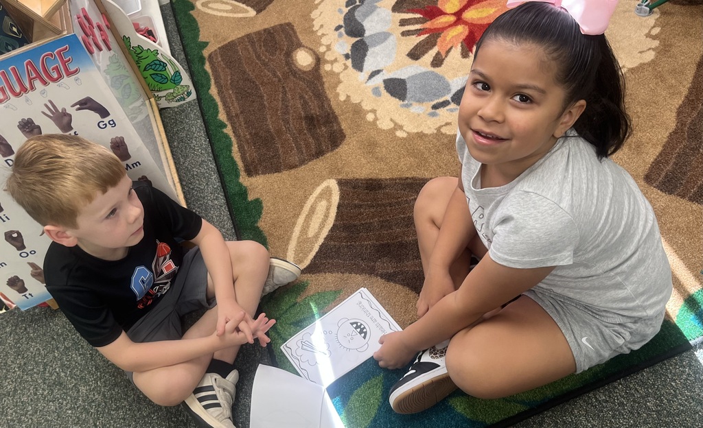 Two children sit on a rug, one holding paper, the other smiling, with a thematic rug in the background.