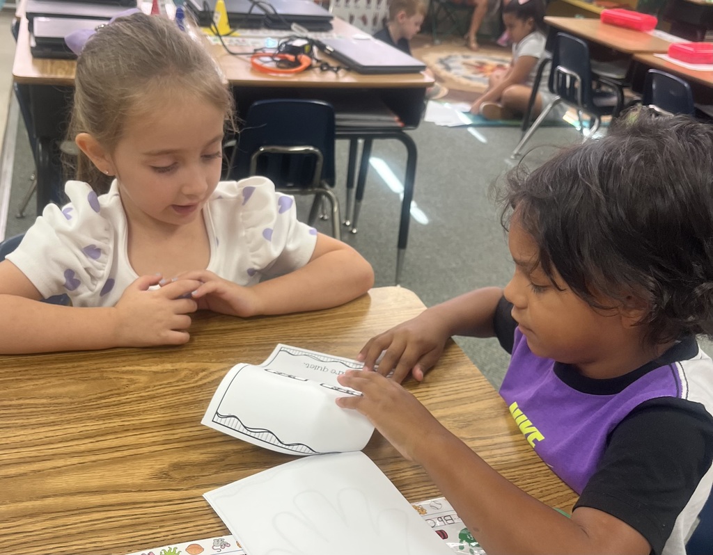Two children sit at a table in a classroom, one pointing at a worksheet. A blurry background shows tables and chairs.