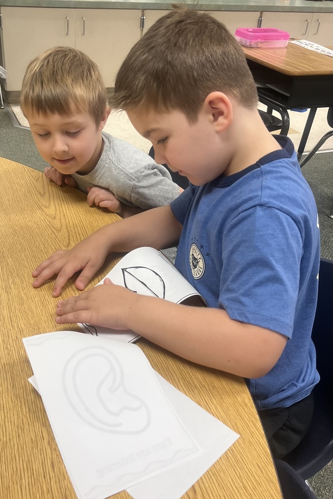 Two boys seated at a desk, one leaning over the other. They have paper with drawn shapes.