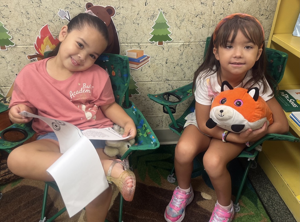 Two young girls in pink shirts and shoes sit in green chairs, smiling, with one holding a paper and the other holding a stuffed animal.