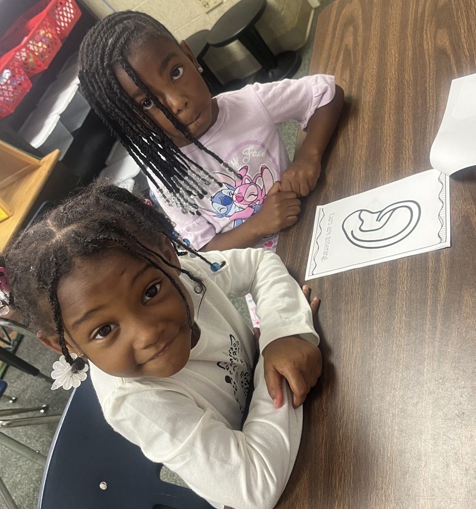 Two young girls sitting at a table, one with braided hair, both focused on a paper with the letter S.