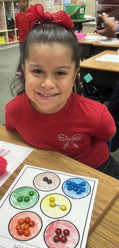 A young girl with a red bow sits at a desk, smiling, and wearing a red shirt with a white bow. A paper with colored candies is on the desk in front of her.