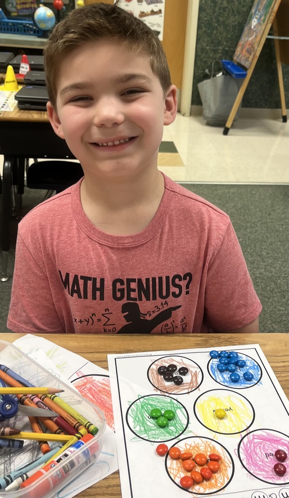 A smiling boy sits at a desk with a red T-shirt. In front of him is a worksheet with colored circles and crayons.