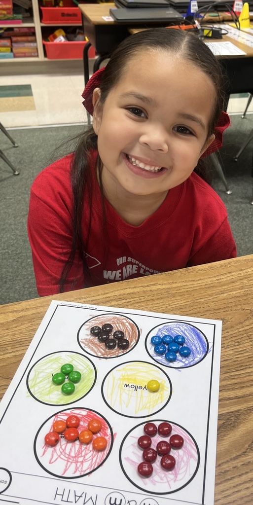 A young girl sits at a table with a colorful counting worksheet, smiling and surrounded by various candies in different colors.