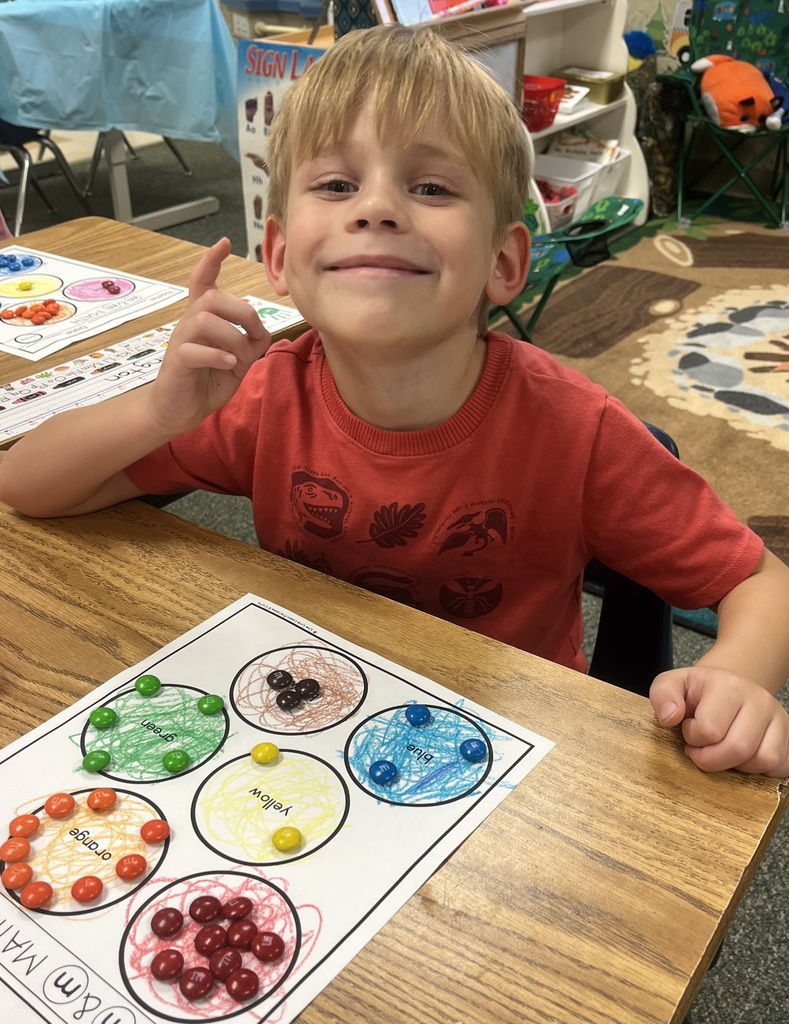Child seated at a table, smiling and pointing to a worksheet with colored dots in circles.