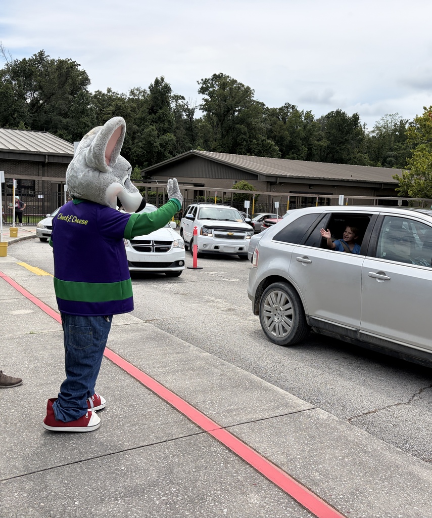 A mascot stands in a parking lot directing traffic. Vehicles, trees, and a building are in the background.