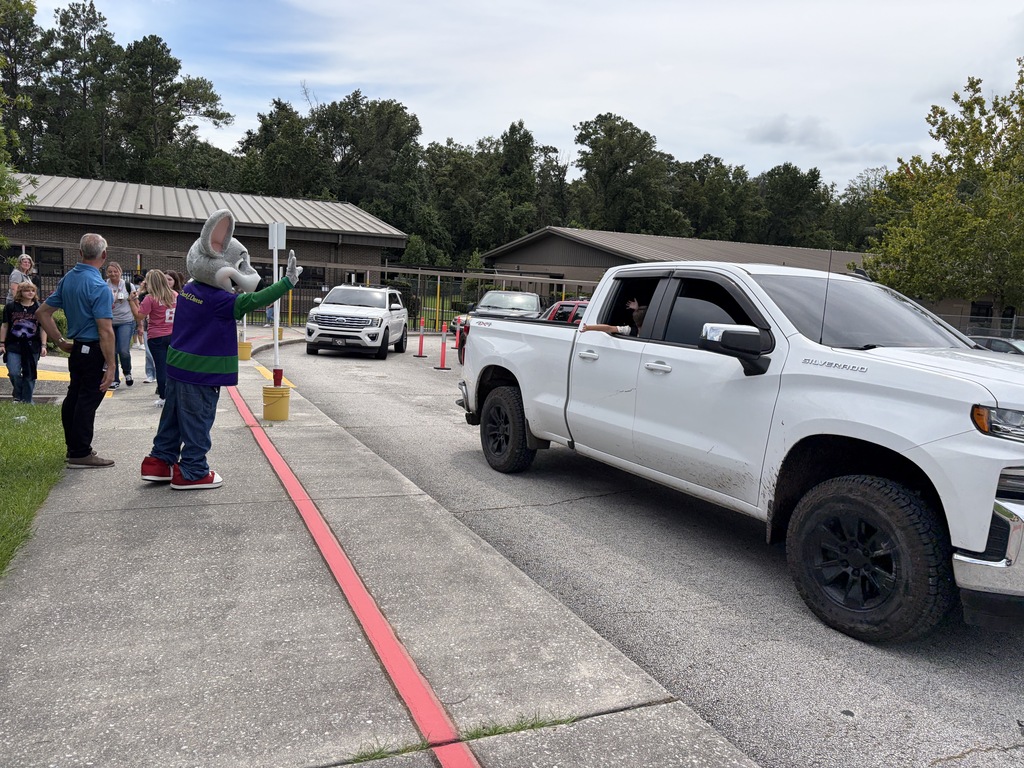 A white pickup truck drives past a person in a mascot costume. A line of people stand near a building.