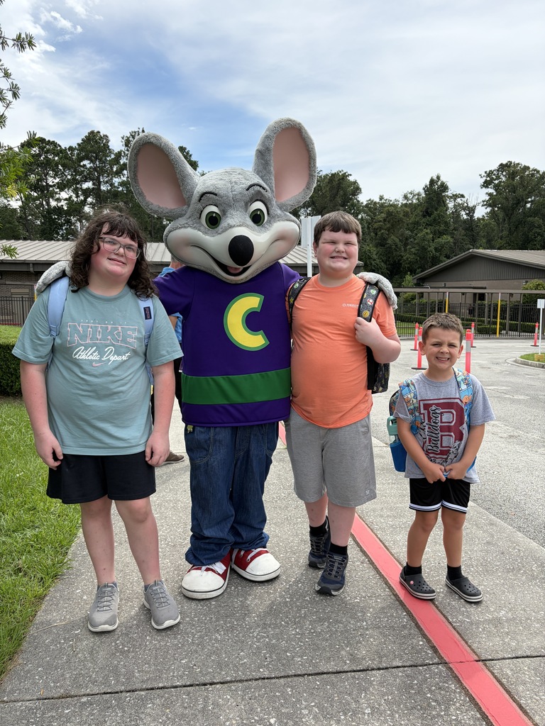 Four children, one in a mouse mascot costume, stand on a sidewalk with grass and buildings in the background.