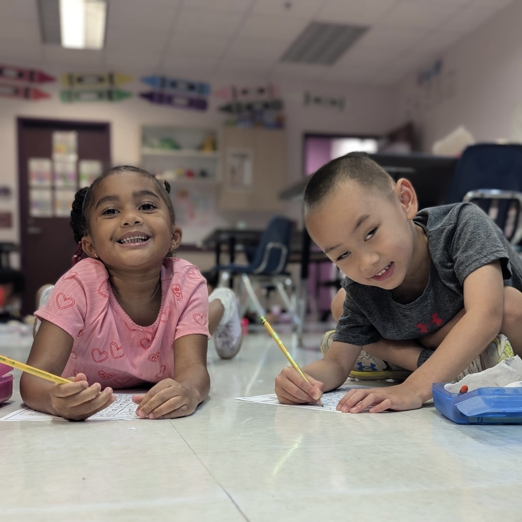 Two children lie on the floor, smiling, and writing on paper with pencils. Background shows a classroom with chairs and a desk.