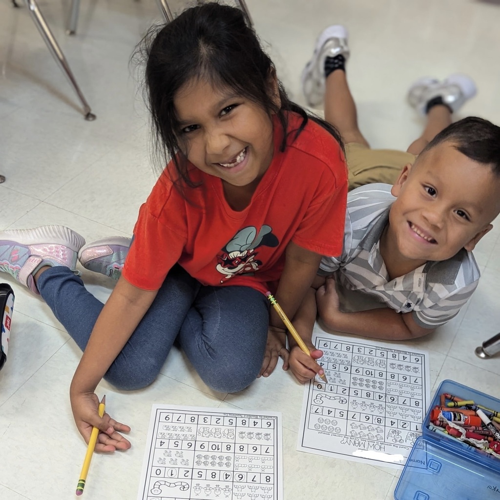Two children, one seated with a pencil, another lying down holding a pencil, smile and appear to work on a worksheet.