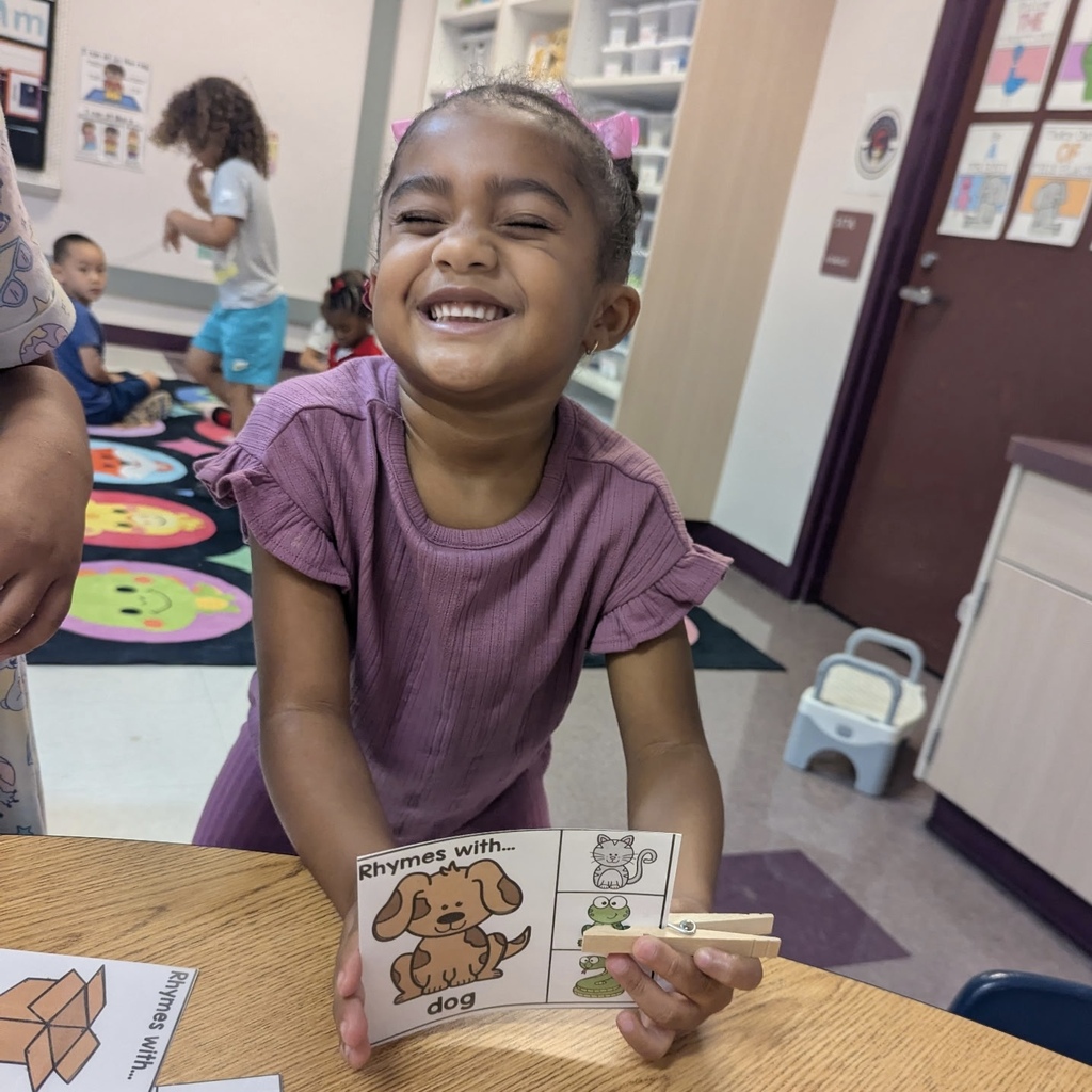 A young girl in purple holds a book with an image of a dog, smiling. She sits at a desk in a classroom with colorful carpet.