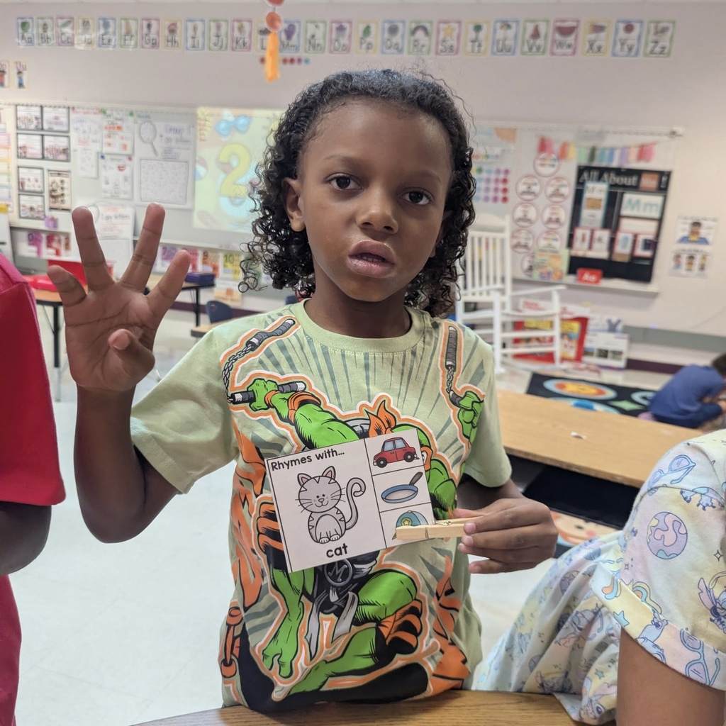 A child in a classroom holds up a card with the word "cat" and makes a peace sign.
