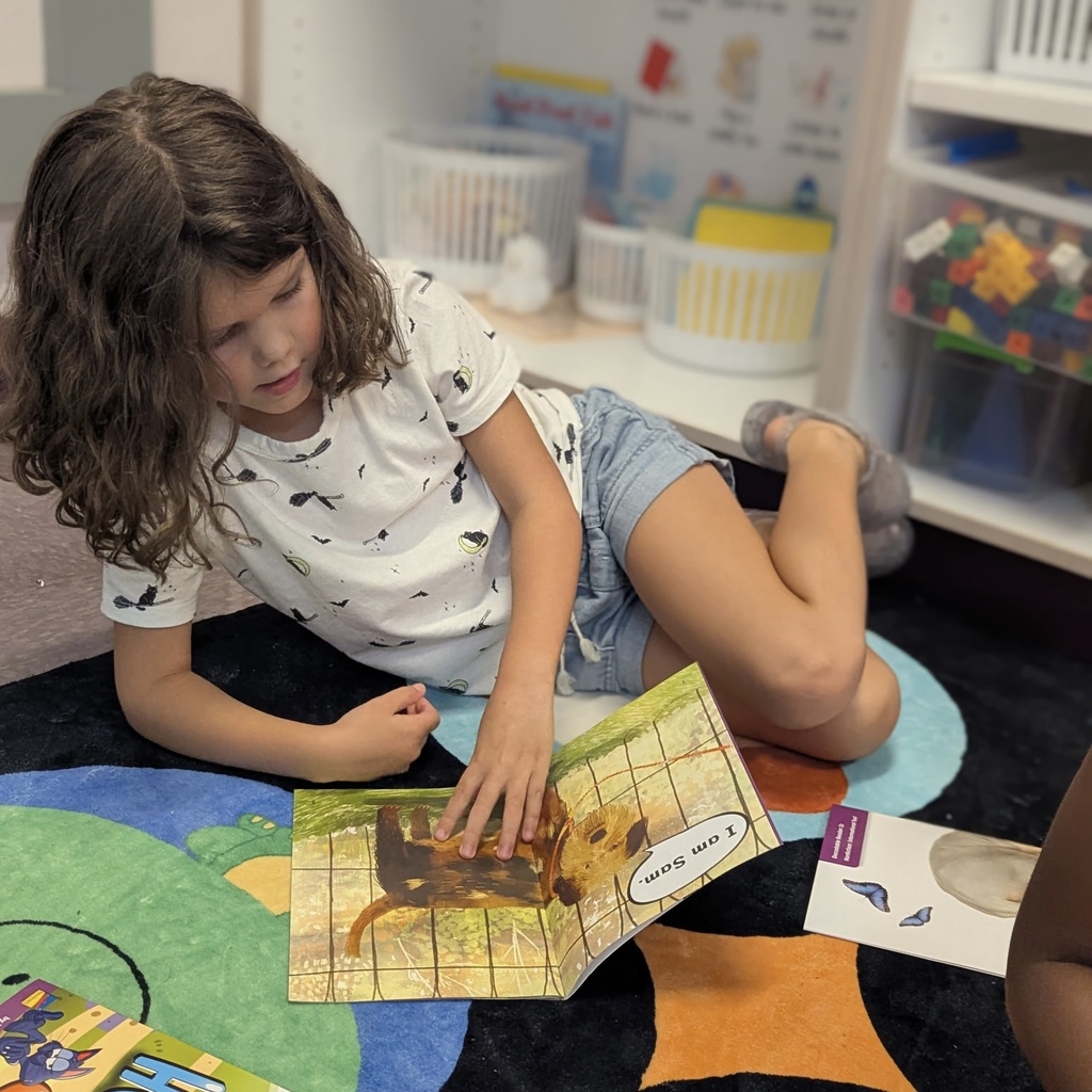 A child sits on a carpeted floor, reading a book with an animal illustration.