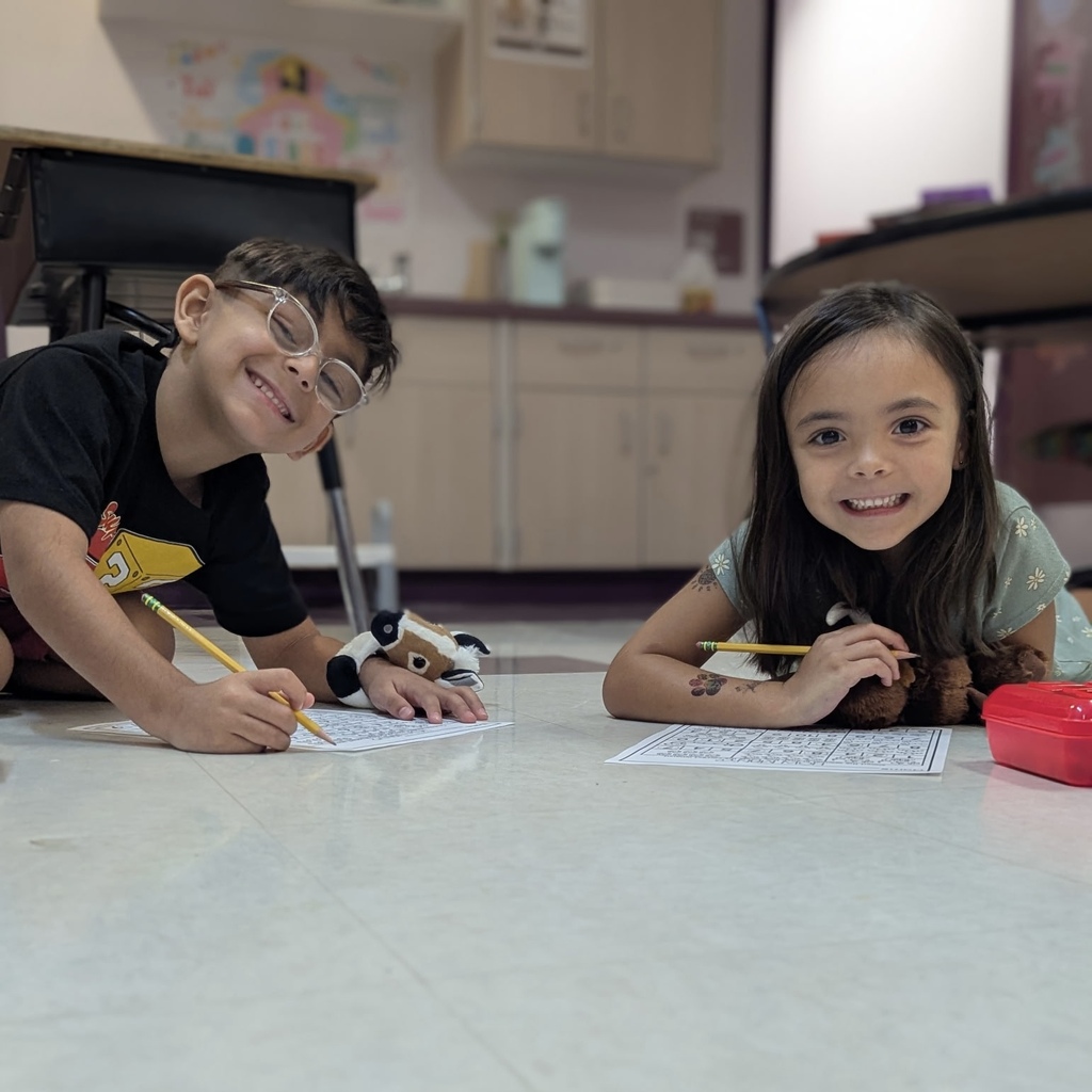 Two children, one boy and one girl, seated on floor with paper and pencils. Boy wears glasses.