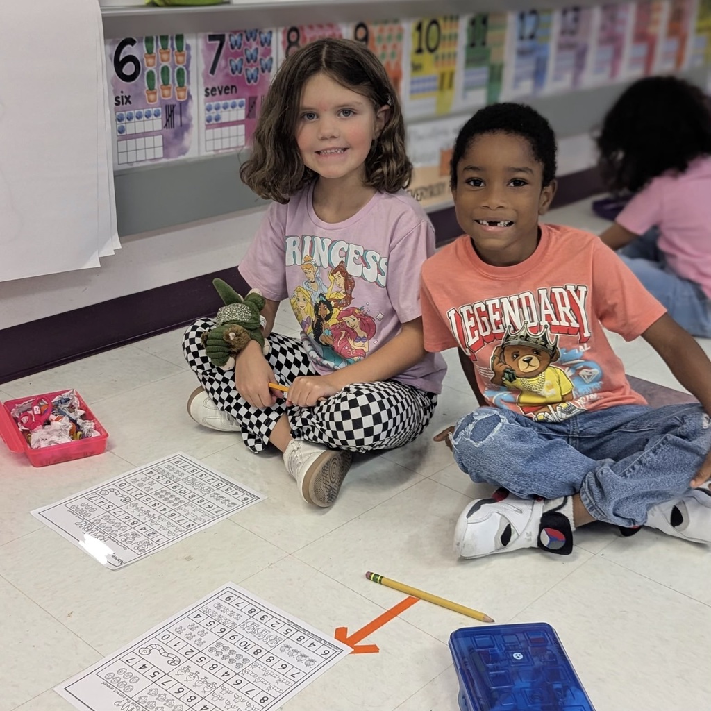 Two children sit on the floor holding pencils, surrounded by papers and a box. One holds a toy.
