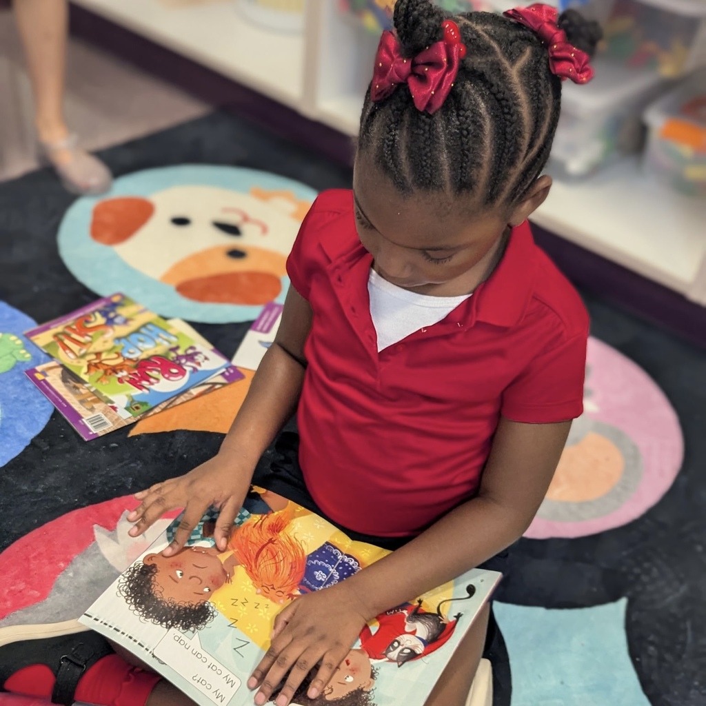 A young girl in a red shirt reads a book with her hands, sitting on a colorful floor mat.