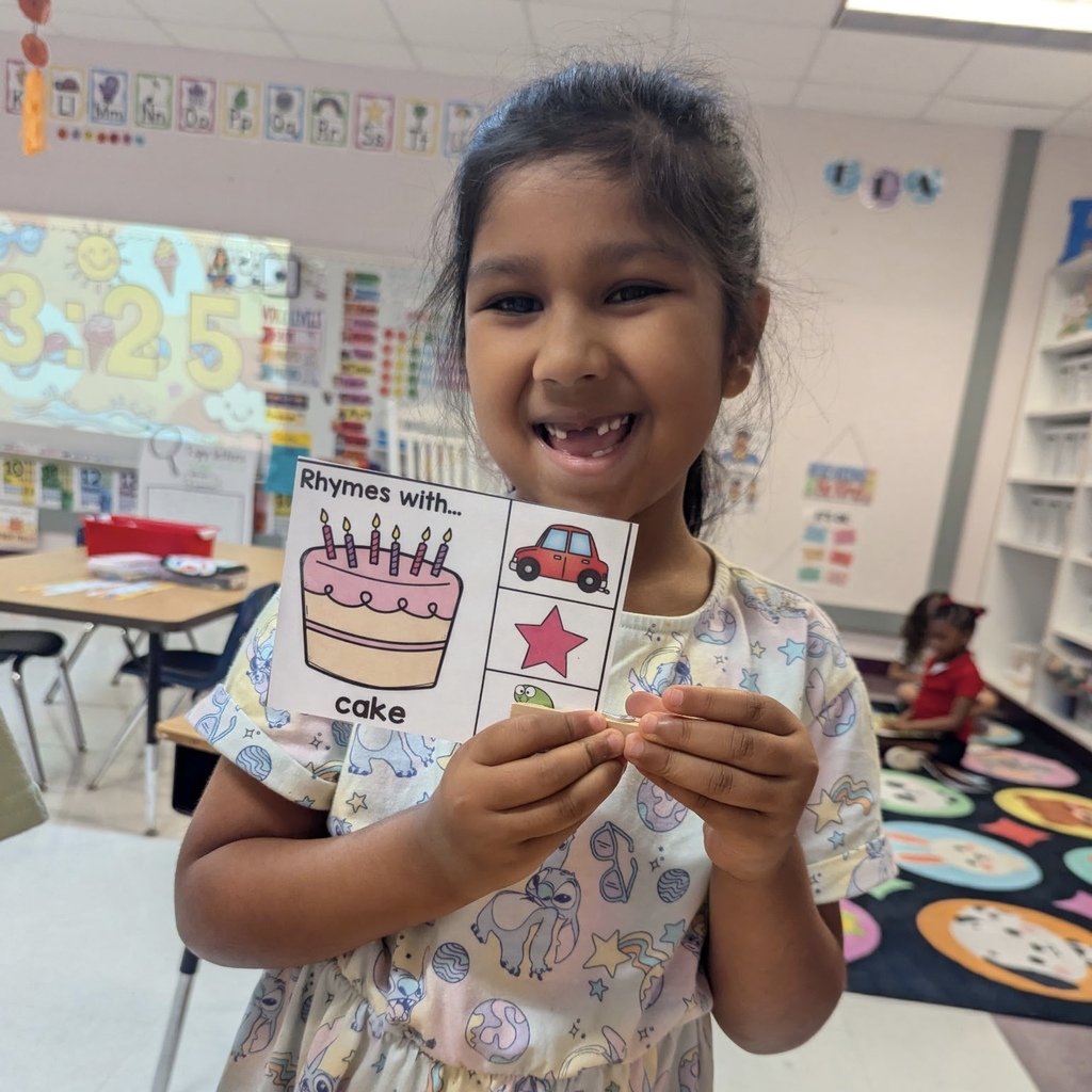 A girl holding a card with a birthday cake and text in a classroom. Other children sit on the floor.