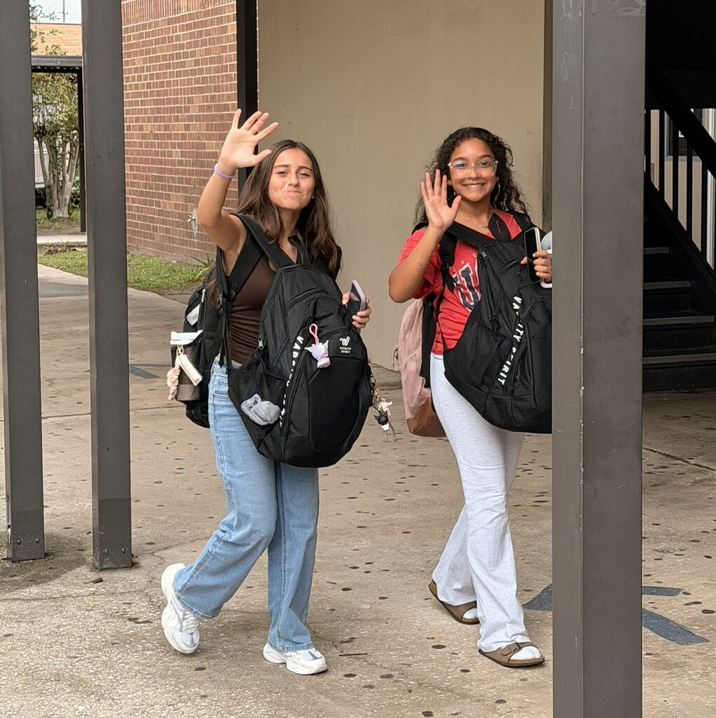 Two girls wave in front of a brick building with a staircase, both wearing backpacks and sneakers.
