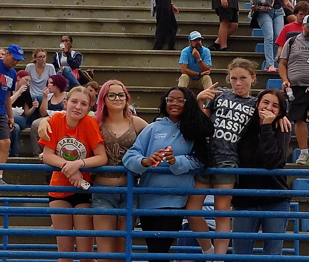 Several girls in a stadium stand by a blue railing. They are looking at a cellphone.