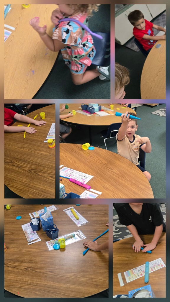 Multiple children are seated at a round table. They are engaged in a classroom activity. Each child holds a stick and colored pieces of paper.
