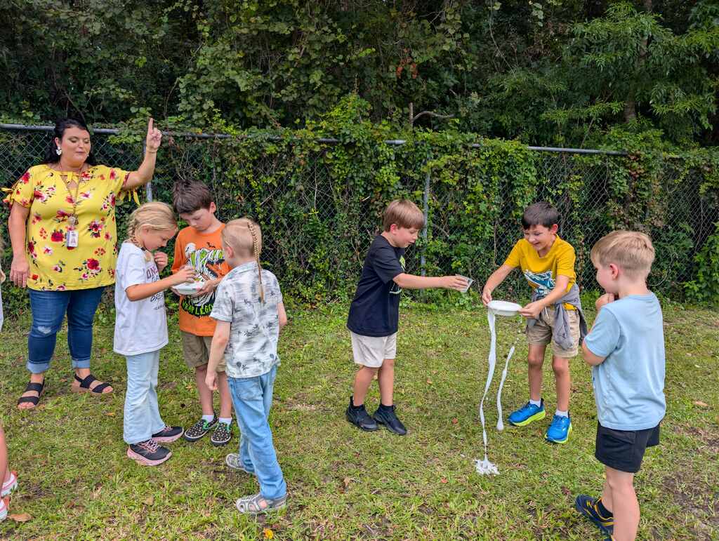 A group of children and an adult hold long pieces of string in a grassy area.