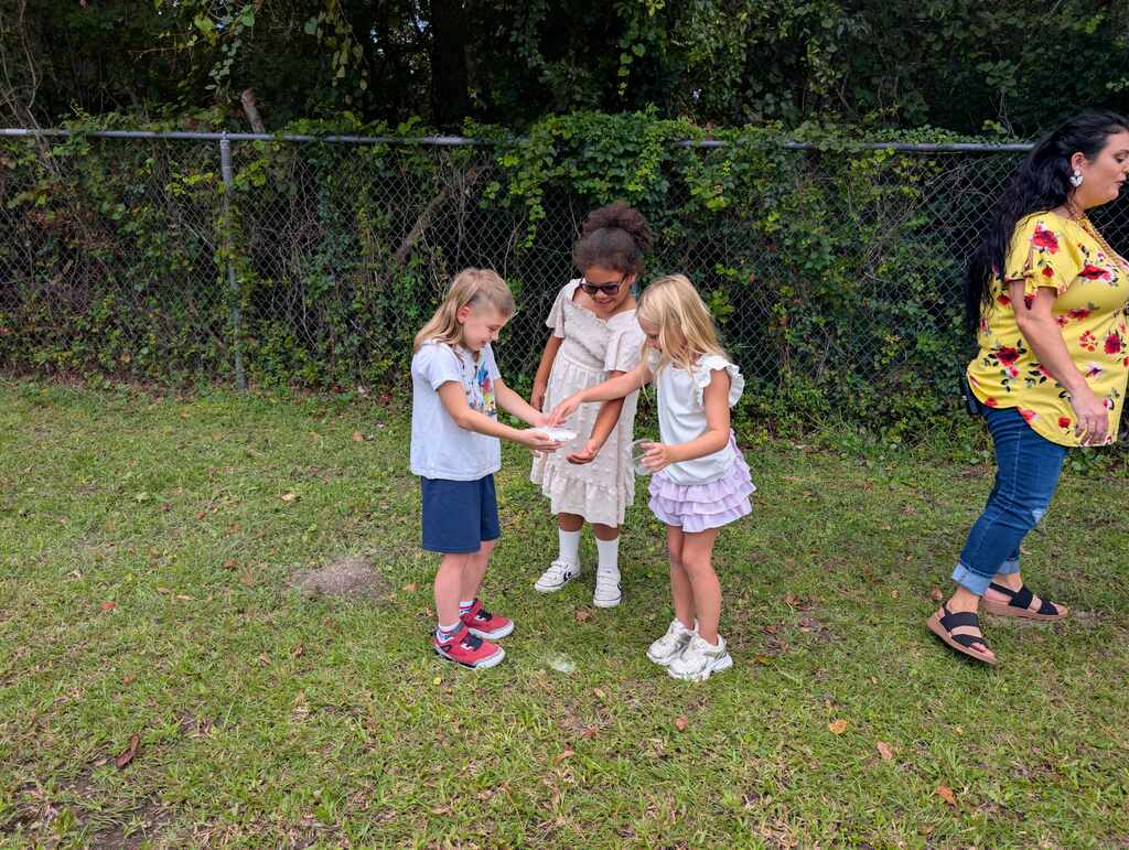 Four children and a woman stand on a grassy field with hands joined, near a chain-link fence.