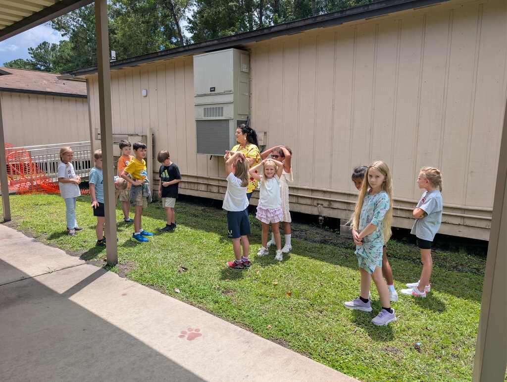 Group of young children in a grassy area, some holding objects, and adults standing behind them. Behind them is a building with a metal fence and an air conditioning unit.