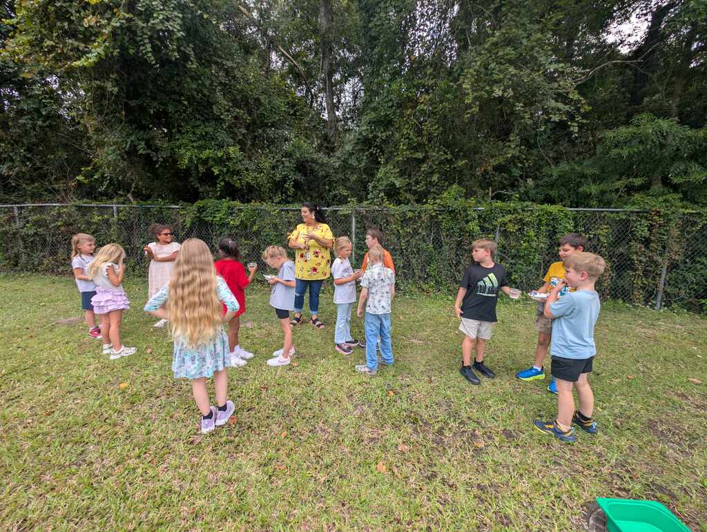 Several children are gathered in a grassy area, some wearing sneakers. A fence and trees are in the background.