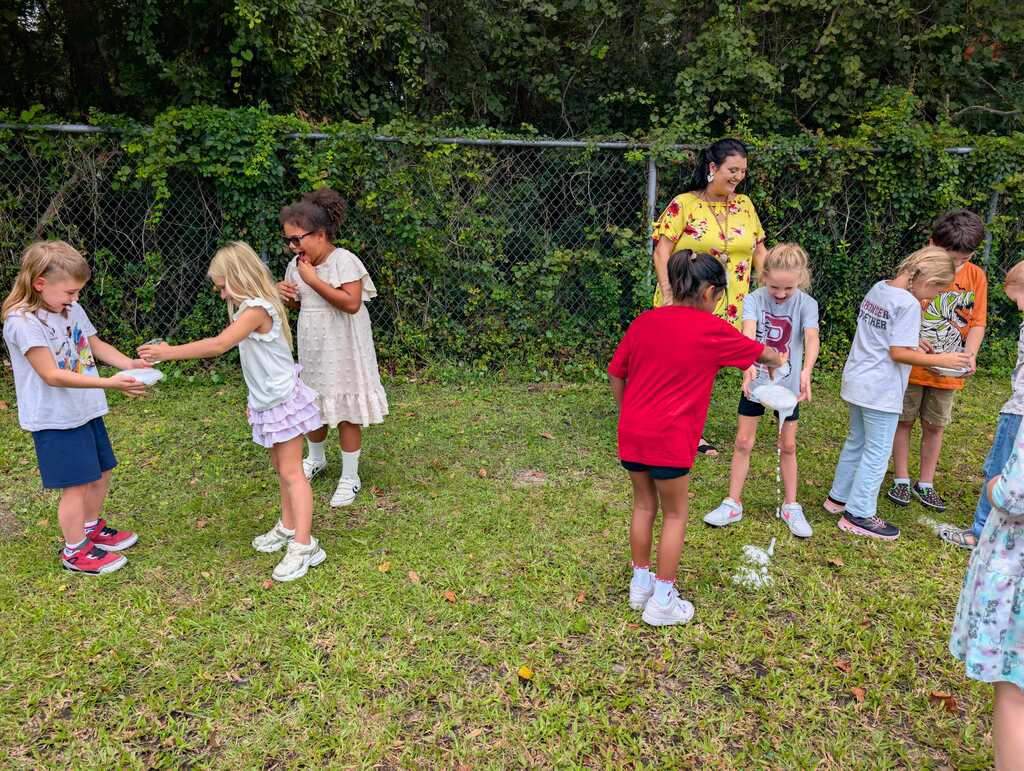 A group of children and adults outdoors in a yard, some holding objects, with a fence and greenery in the background.