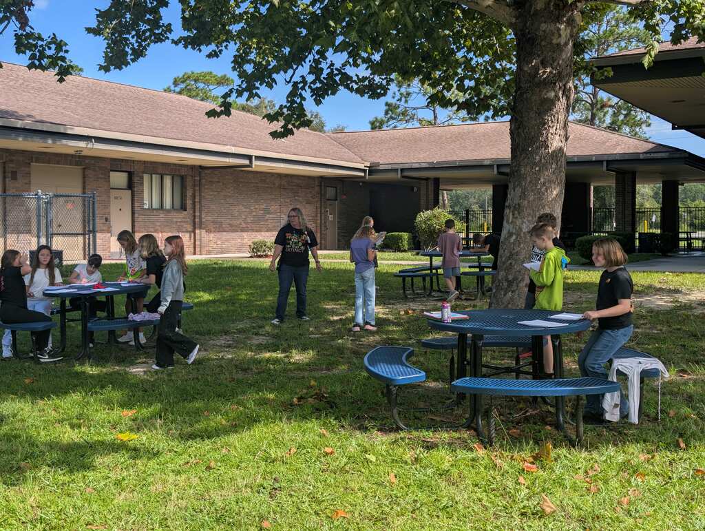 People gather in a grassy area with picnic tables in front of a building with a brown roof.