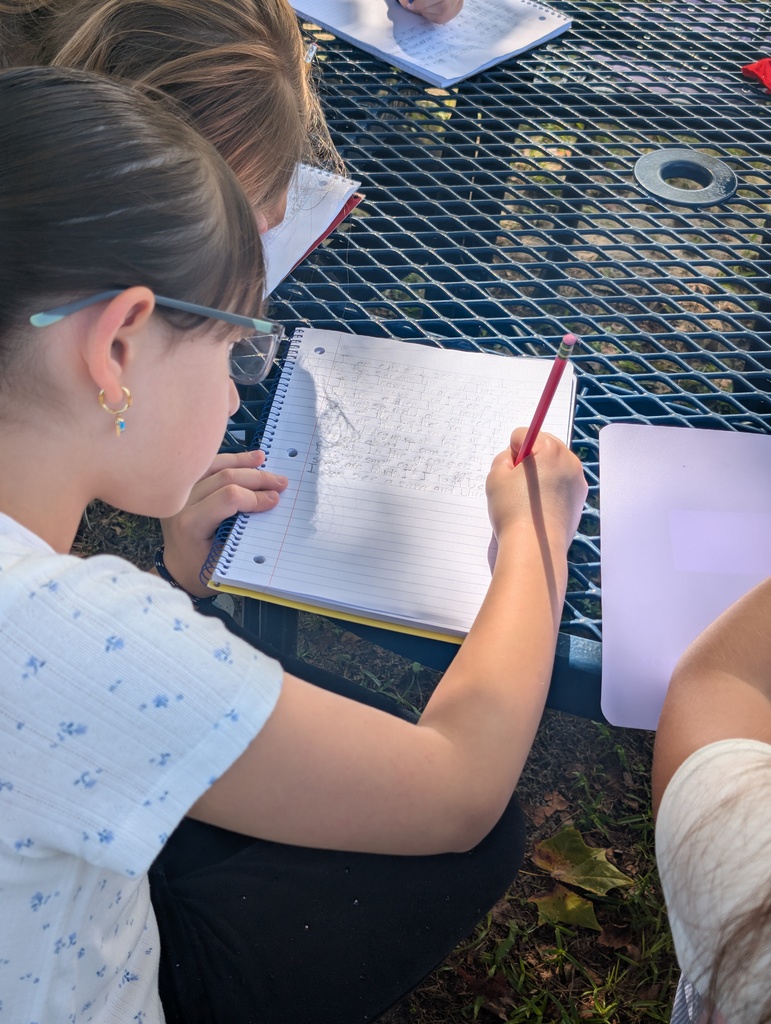 Two people are seated around a table, each holding a pen and notebook. One person is drawing.