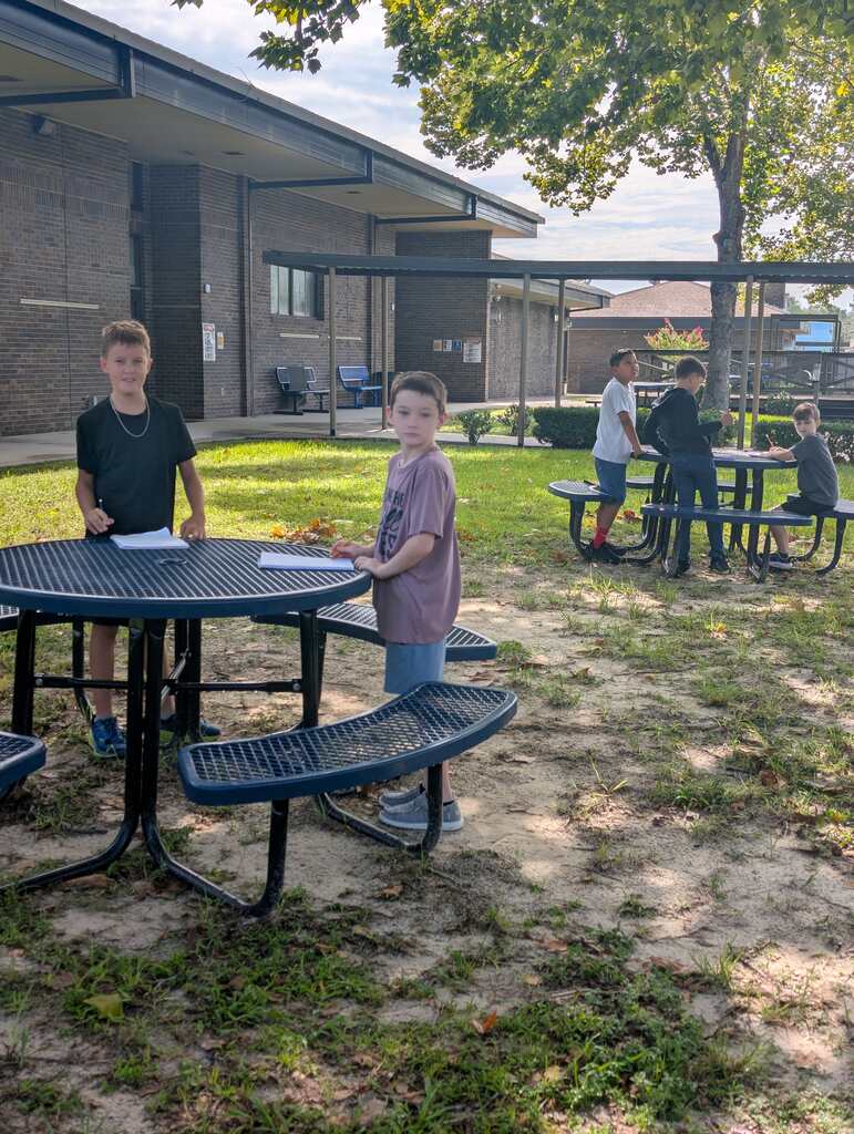 A group of individuals outside a building. Two boys stand by a table, and others sit at tables.