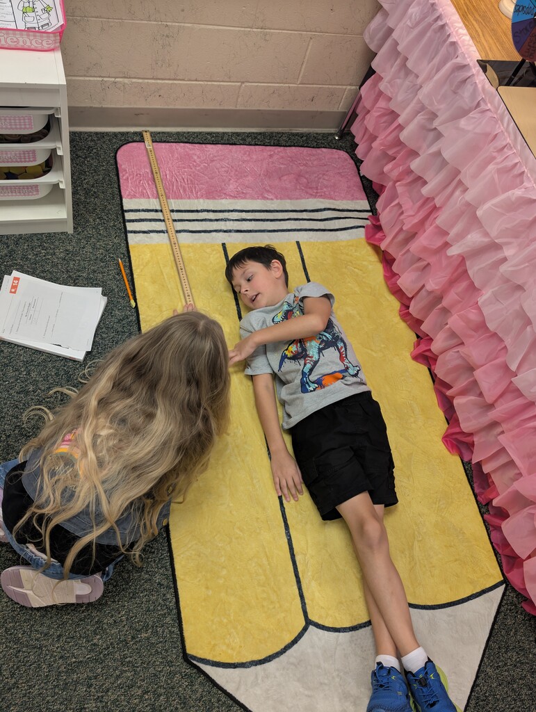 Child measuring pencil rug, one child lying down, another crouching, with a ruler. Background includes classroom furniture.