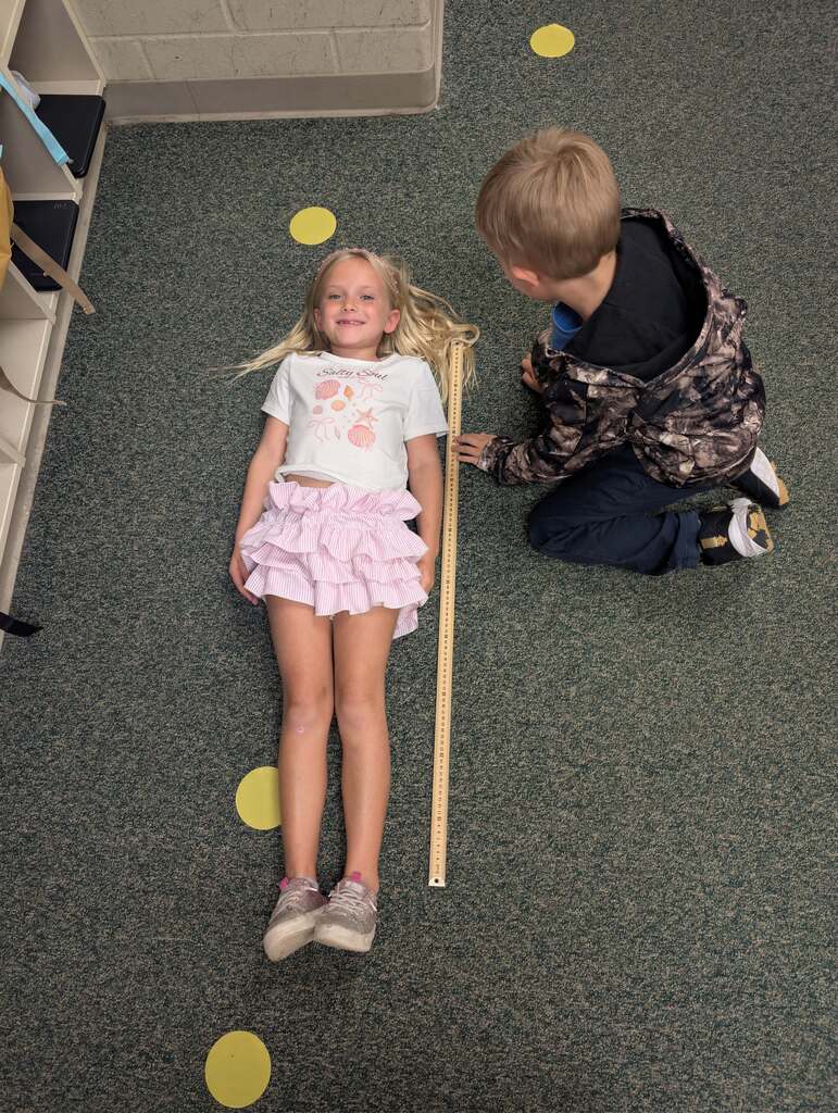 A boy measures a girl's height with a ruler on a carpeted floor. Yellow circles are scattered on the floor.