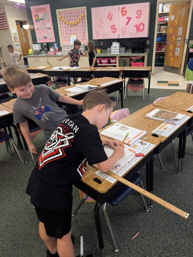 Two boys working at desks in a classroom with other students, a whiteboard, and number charts.