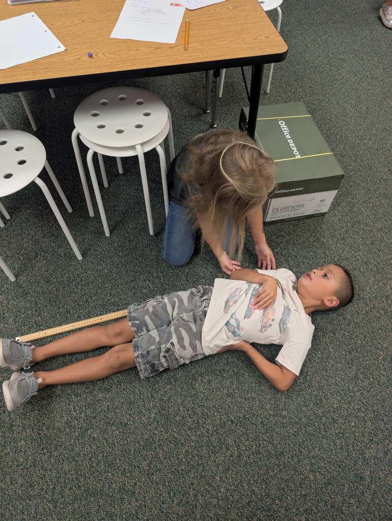 A person assists a child lying on the floor, holding a stick, in a room with stools and a desk.