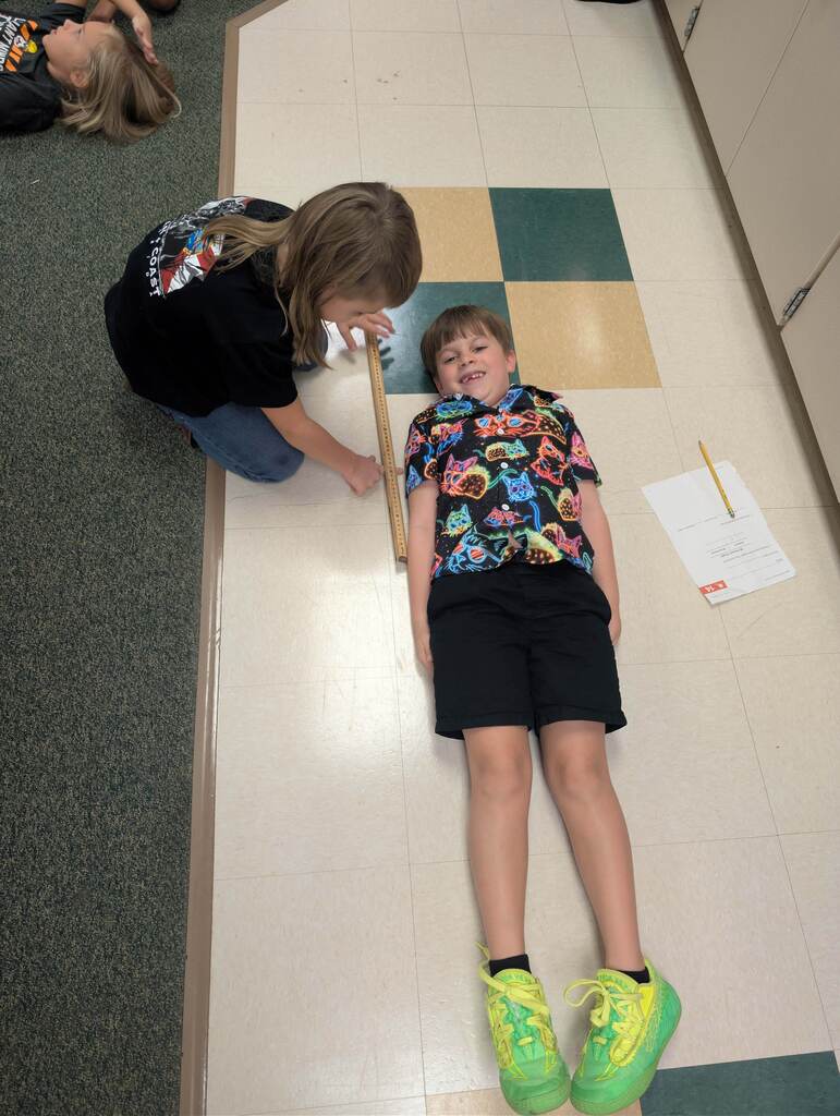 A child lies on the floor as another child measures their height with a stick. White floor tiles and gray carpet visible.