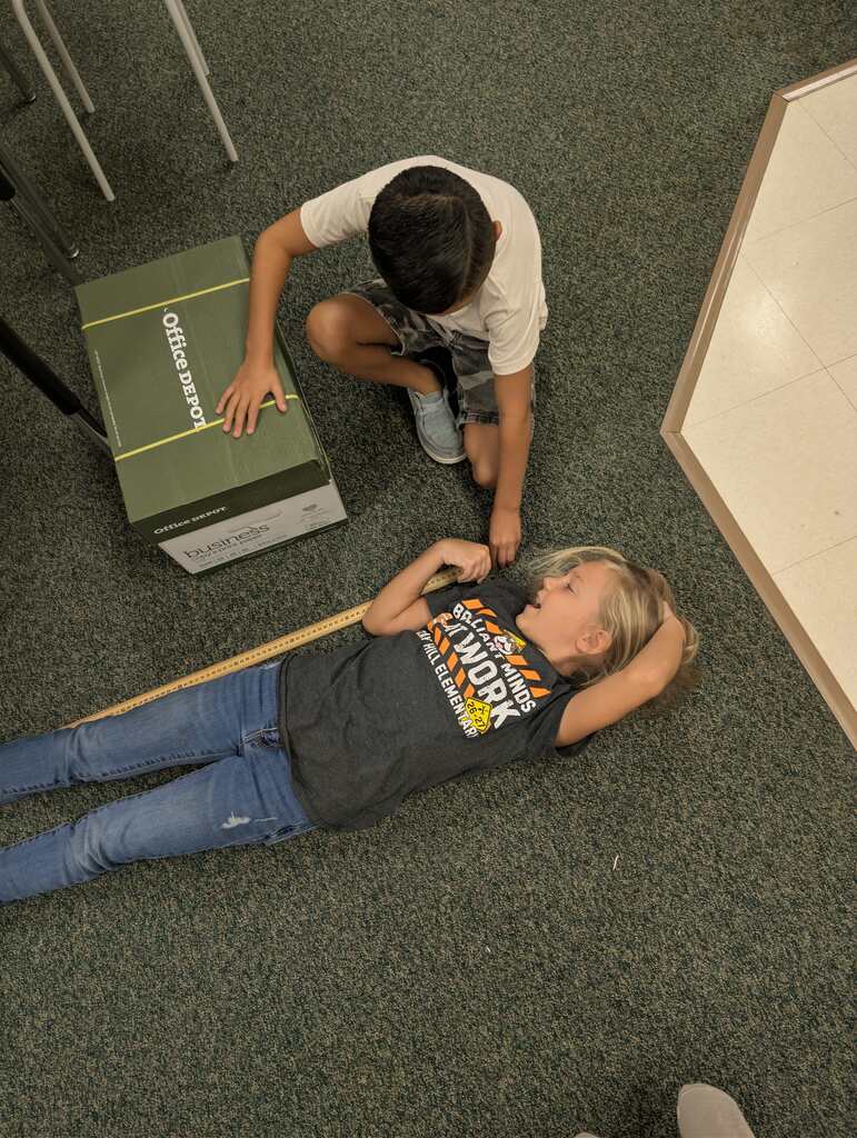 Two children in a room. One is kneeling, opening a box. The other lies on the floor, looking at the box.