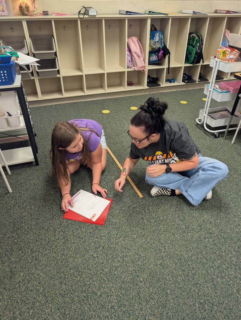 Two individuals in a classroom setting, kneeling on a carpeted floor, with one measuring a red folder.