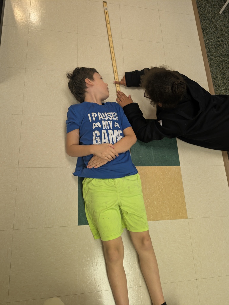 A boy lies on a tiled floor while a person takes his measurement with a ruler.