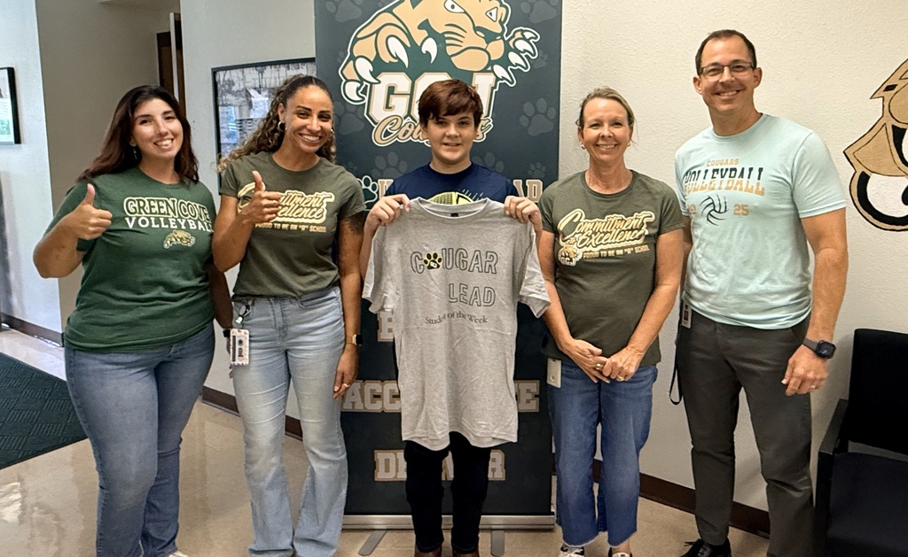 Five adults stand with a white T-shirt between them. Three wear green, and two wear light blue. They smile for a photo.