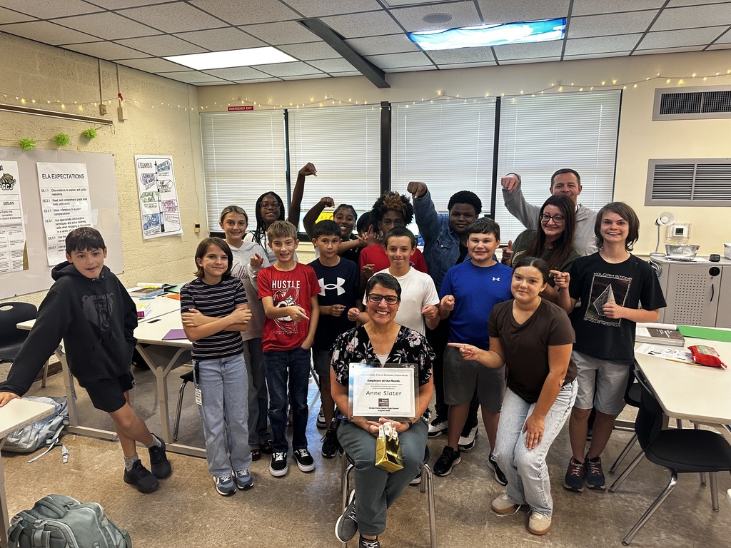 A classroom with kids, a woman in glasses, and a certificate. A white table is in front of them.
