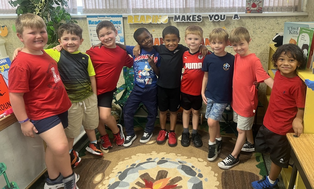 Children stand in a circle in a classroom, wearing various colored t-shirts and shorts. Some have sneakers.