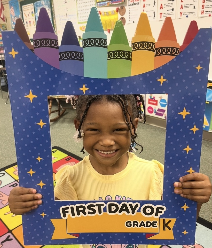 Girl in a classroom holds a frame with crayons on top. Frame says "First Day of Grade K".