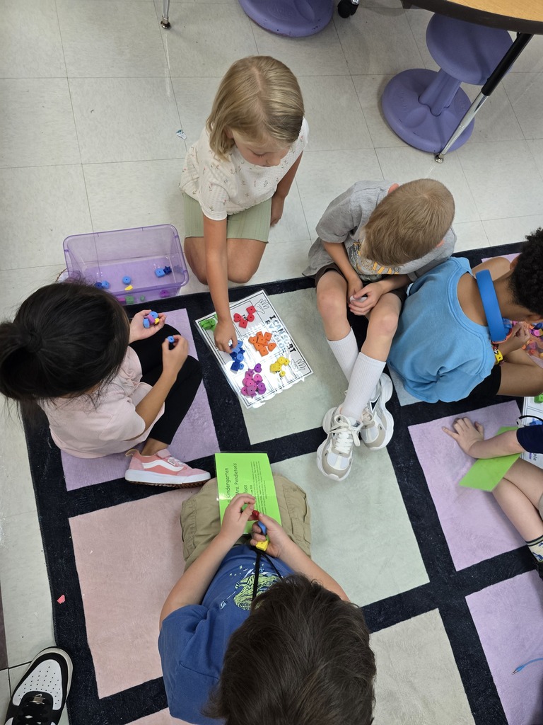 A group of children, all engaged in a classroom activity, sit on a colorful mat. Each child holds a piece of paper or a small object.