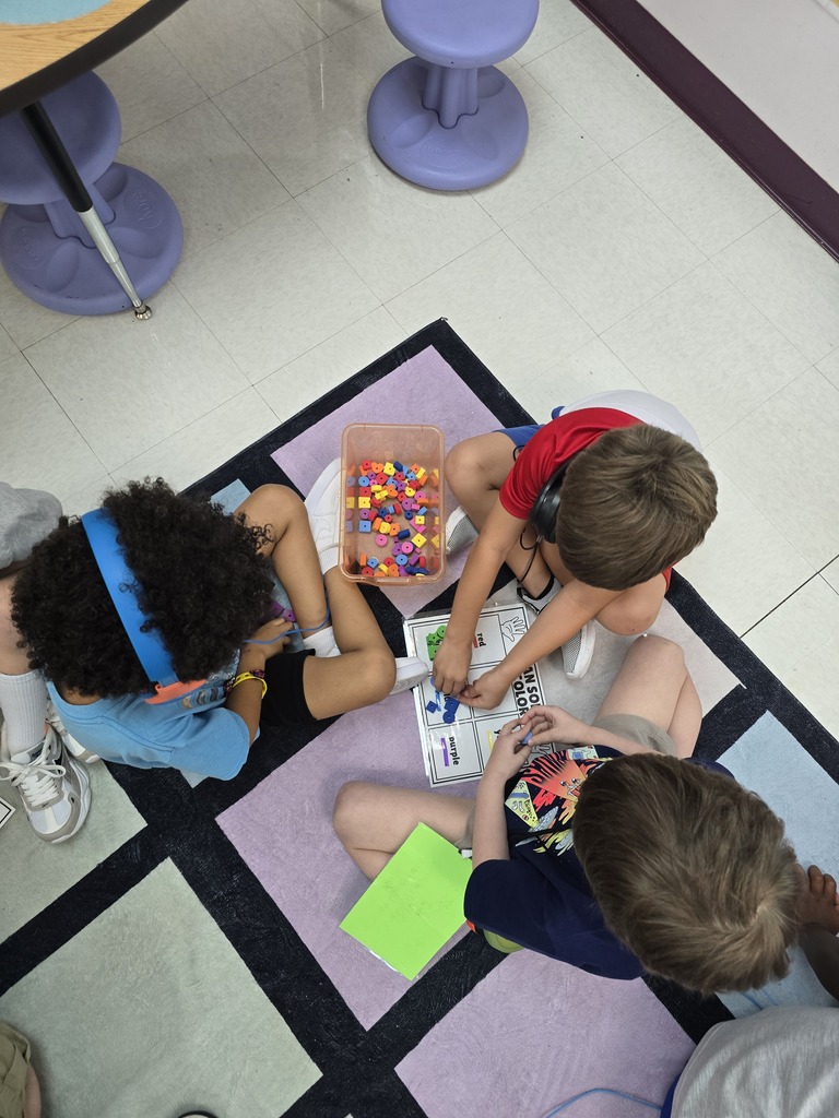 Three children sit in a circle on a rug. They work on a project, holding colorful beads and papers.