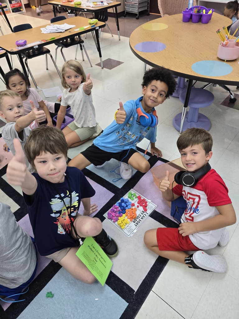 A classroom with children sitting on the floor. Several children are giving a thumbs up. A table with chairs is in the background.