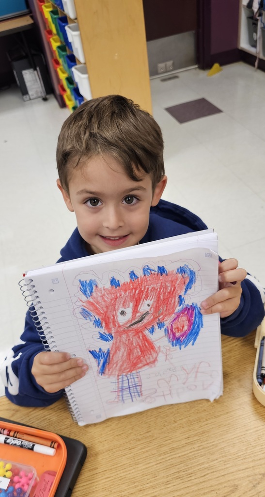 Child holding a notebook with a drawing of a red creature. The child smiles, wearing a blue jacket.